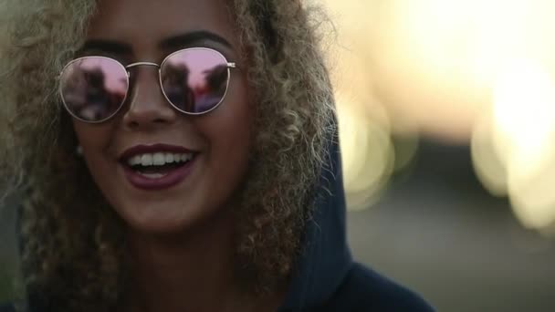 Hipster tjej med lockigt blont hår och speglade solglasögon tittar bort och ler. — Stockvideo
