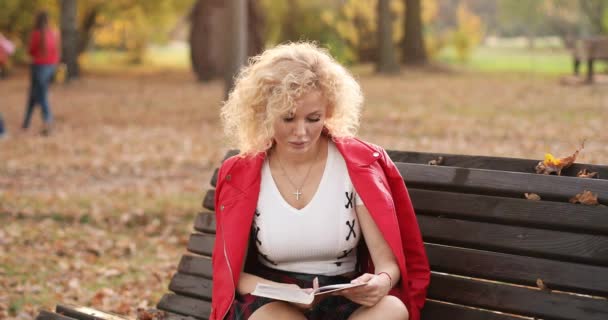 Jolie jeune femme lisant un livre sur le banc à l'extérieur au coucher du soleil . — Video