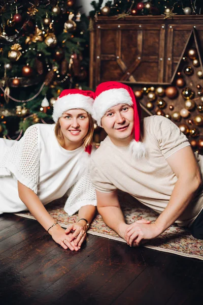 Smiling couple in red festive hats shoulder to shoulder on the floor. — Stock Photo, Image