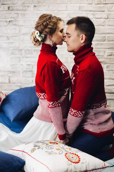 Pareja en jerséis rojos de Navidad besándose rodeada de almohadas . — Foto de Stock