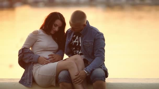 Беременные родители сидят на пирсе озера на закате и обнимают желудок . — стоковое видео