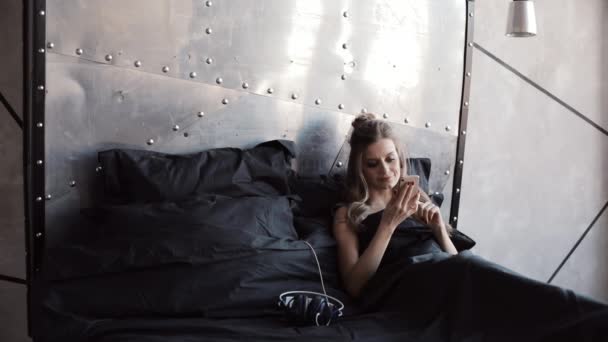 Hermosa rubia revisando su teléfono móvil sentada en la cama bajo una manta negra . — Vídeo de stock