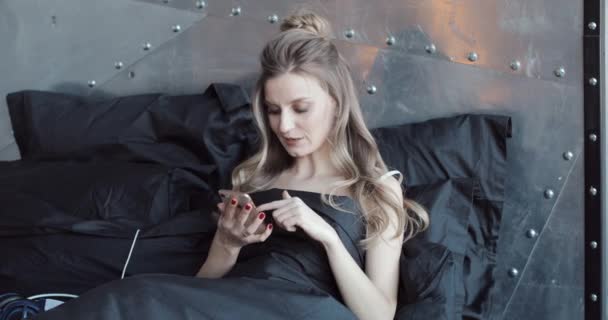 Πανέμορφο blondie ελέγχοντας το κινητό της τηλέφωνο κάθεται στο κρεβάτι κάτω από κουβέρτα μαύρη. — Αρχείο Βίντεο