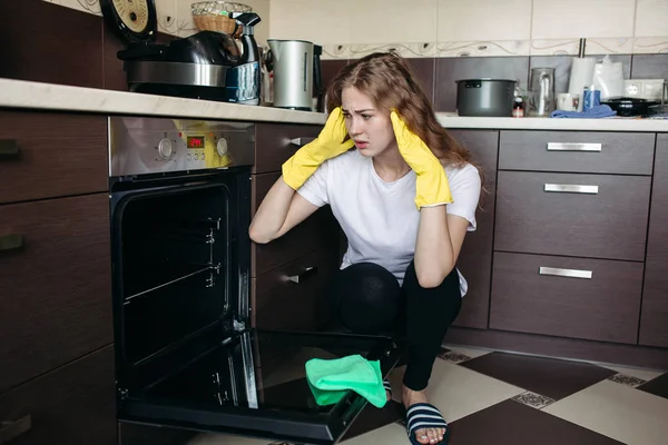 Mujer en guantes protectores amarillos buscando horno sucio en la cocina casera . — Foto de Stock