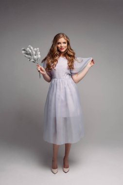 Güzel uzun elbiseli dal köknar ağaçlarının tutmak ve kamera Studio bakarak çekici modelinin önden görünüm. Genç ve güzel sarışın makyaj ile ve uzun gri izole arka plan üzerinde poz bukleler.