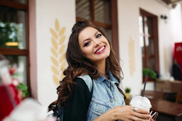 Bella ragazza con bruna capelli ondulati in gilet di denim con bevanda nel caffè . — Foto Stock