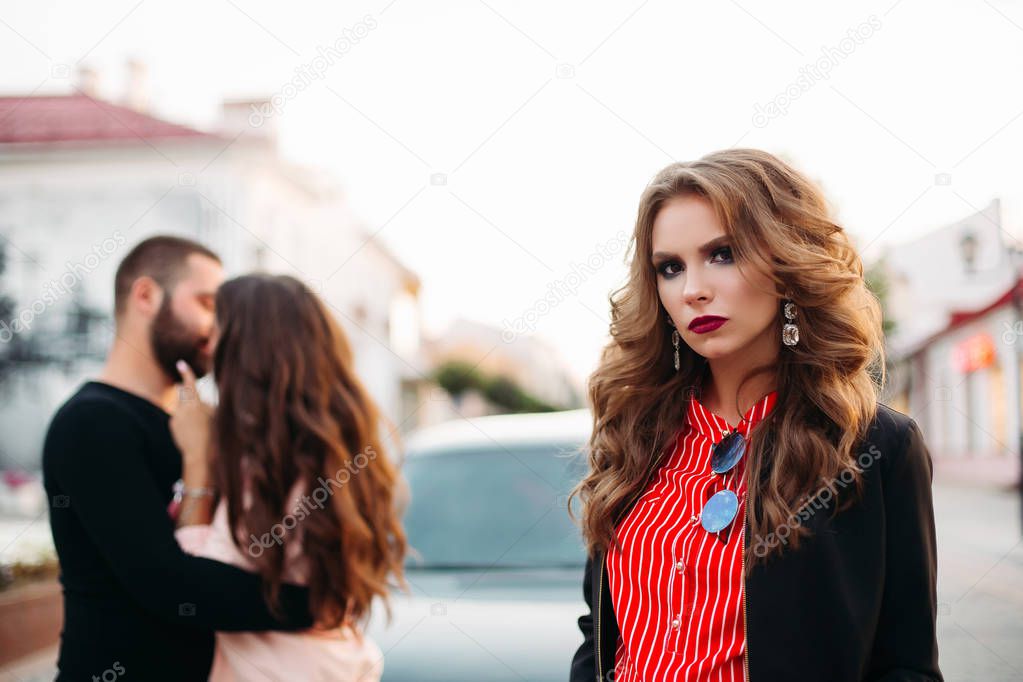 Portrait of beautiful woman with make up looking at camera sadly against unfocused kissing couple.