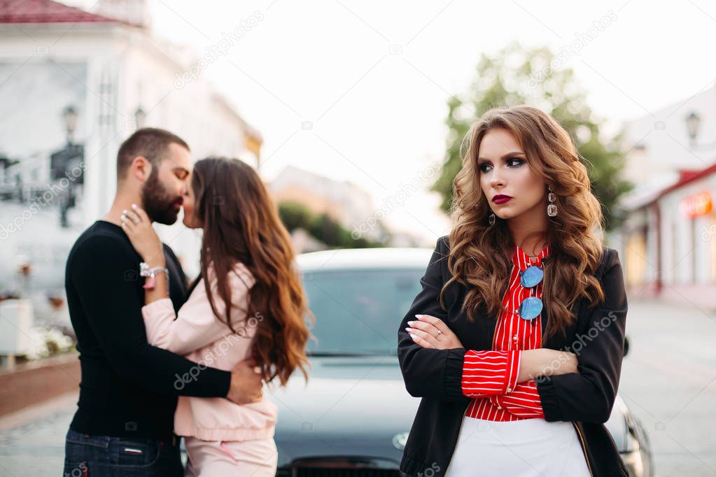 Sad beautiful woman with folded arms in fashionable clothes against kissing couple.
