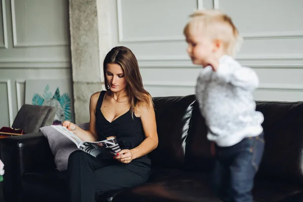 Matka čtení časopisu, zatímco její syn skákat na gauči. — Stock fotografie