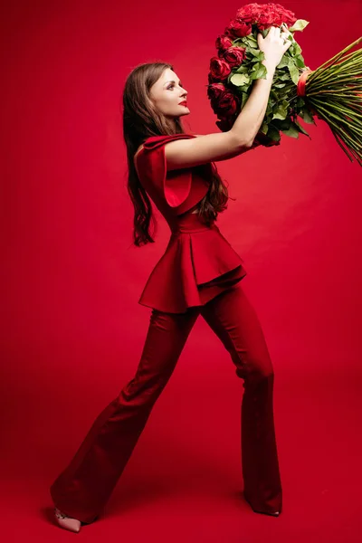 Jovem morena feliz em vestido vermelho segurando grande monte de rosas vermelhas . — Fotografia de Stock