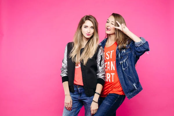 La imagen primaveral de dos chicas positivas con gafas, novias con pelo liso que se abrazan, miran a la cámara. Una chica una chaqueta de mezclilla pone cuernos de novia. En ropa de estilo cotidiano y gafas . — Foto de Stock