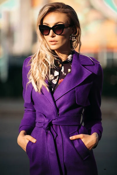 Молодая красивая стильная женщина в солнцезащитных очках и фиолетовом пальто позирует на улице . — стоковое фото