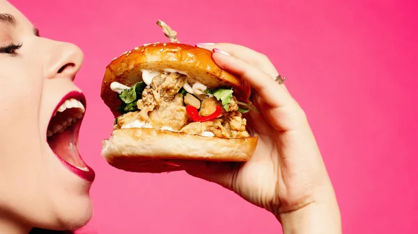 Woman eating sandwich. The happy girl is eating a hamburger. She opened her mouth, holding a hamburger in her right hand and looking at it appetizingly. A student with a meal on a pink background,