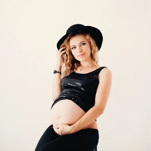 Piękna kobieta w ciąży z nagi brzuch, obejmując ją patrząc na kamery, noszenie czapki i góry. — Zdjęcie stockowe