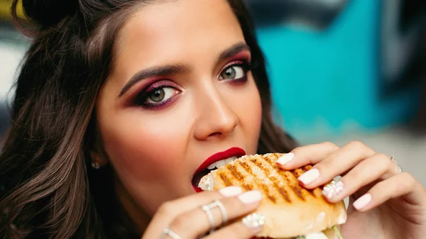 Dziewczyna z jasnym makijażu biorąc gryza hamburgera. — Zdjęcie stockowe