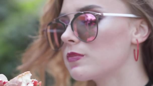 Menina adolescente em óculos de sol elegantes com lábios brilhantes desfrutando de delicioso hambúrguer . — Vídeo de Stock