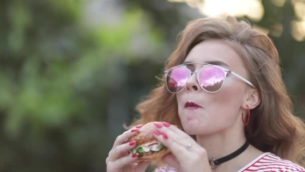 Teen κορίτσι σε μοντέρνα γυαλιά ηλίου με τα έντονα χείλη, απολαμβάνοντας τα νόστιμα burger. — Αρχείο Βίντεο