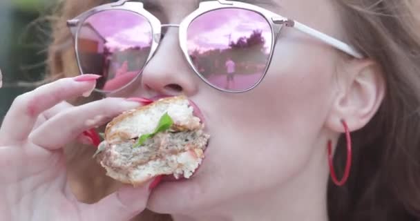Teen dziewczyna w stylowych okularów przeciwsłonecznych z jasne usta ciesząc się smacznego hamburgera. — Wideo stockowe