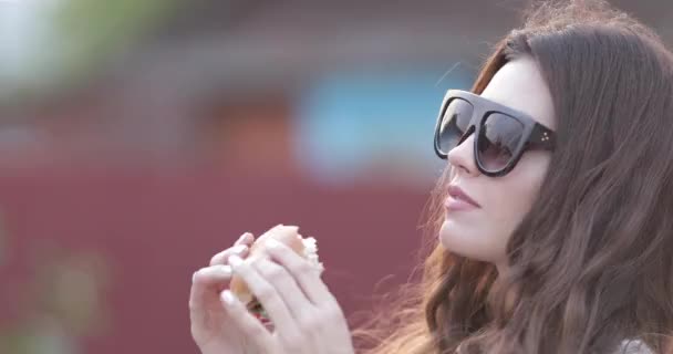 Teen κορίτσι σε μοντέρνα γυαλιά ηλίου με τα έντονα χείλη, απολαμβάνοντας τα νόστιμα burger. — Αρχείο Βίντεο