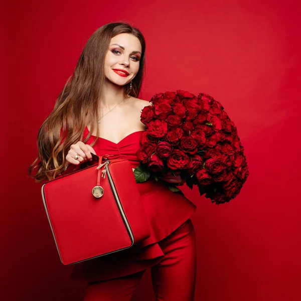 Elegante hermosa morena en rojo con rosas rojas y bolsa roja . — Foto de Stock