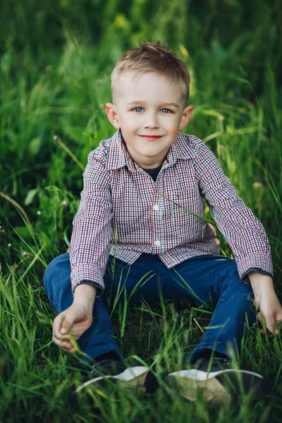 Portret van stijlvolle blond jongetje zitten in park onder gras. — Stockfoto