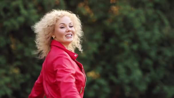 Η όμορφη ξανθιά γυναίκα με σγουρά μαλλιά σε κόκκινο μπουφάν στρίβοντας στο πάρκο. — Αρχείο Βίντεο