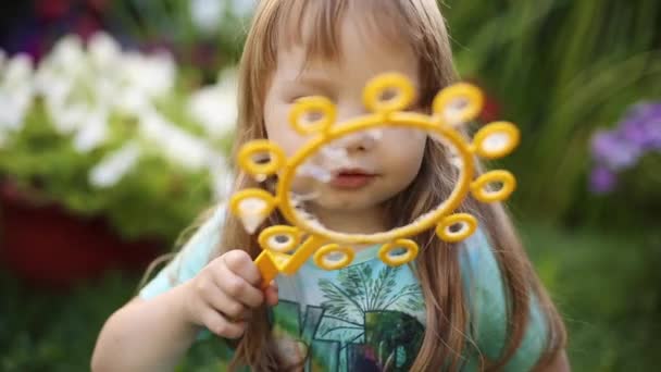 Супер милая светловолосая маленькая девочка в футболке, пускающая мыльные пузыри на траву. . — стоковое видео
