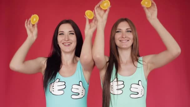 Hermanas felices se meten y juegan con naranja. Vídeo promocional brillante. Morenas sobre fondo rojo con naranjas — Vídeos de Stock