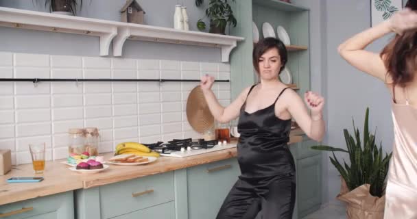 Twee zwangere dames in pyjama dansen en eten cupcakes in de keuken. — Stockvideo