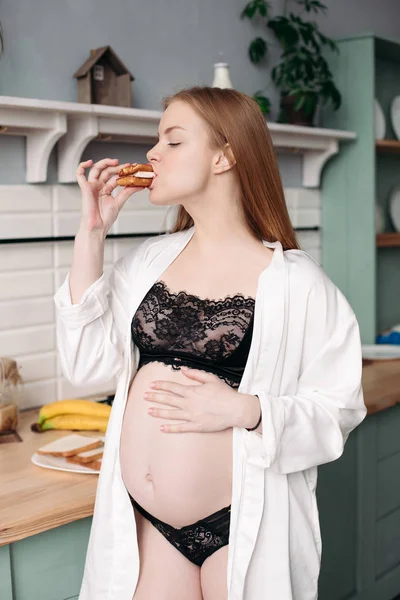Frumos femeie gravidă tânără în lenjerie de dantelă neagră și halat alb care mănâncă micul dejun . — Fotografie, imagine de stoc