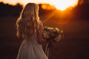 Tanınmayan sarışın gelin uzağa bakıyor ve gün batımında düğün boquet tutarak.