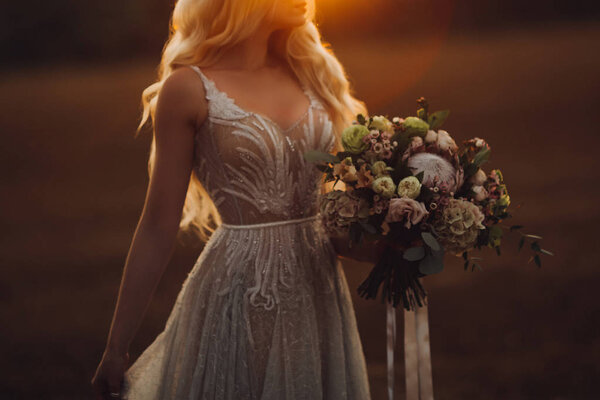 Crop of beautiful long haired blondie bride wearing elegant ivory lace wedding dress, holding by hands and looking at big colorful wedding boquet. Walking in field at sunset time.