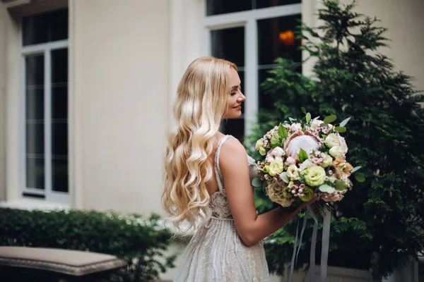 Splendida sposa con mazzo di fiori nuziali in posa all'aperto, contro casa di lusso . — Foto Stock