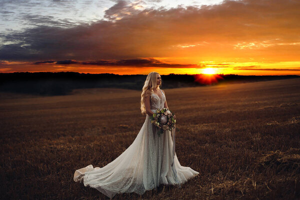 Panoramic view of beautiful bride with wavy hair, wearing in volumed wedding dress looking at side. Pretty blonde woman holding colorful wedding bouquet in hands. Posing at field at sunset time.