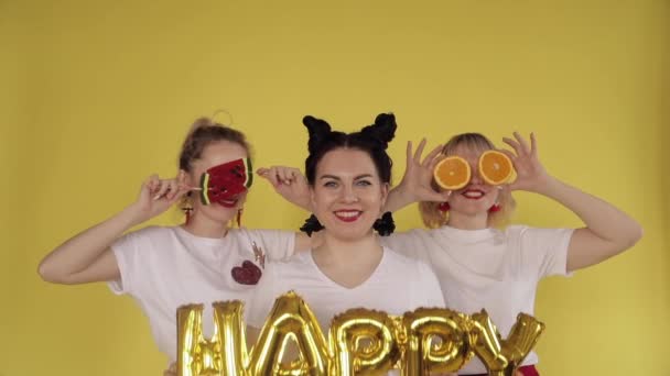 Three girls having fun on yellow background. Chewing gum, playing with candies and fruit. — Stock Video