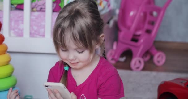 Χαριτωμένο κορίτσι παίζει παιχνίδι στο τηλέφωνο και χαμογελά στο σπίτι — Αρχείο Βίντεο