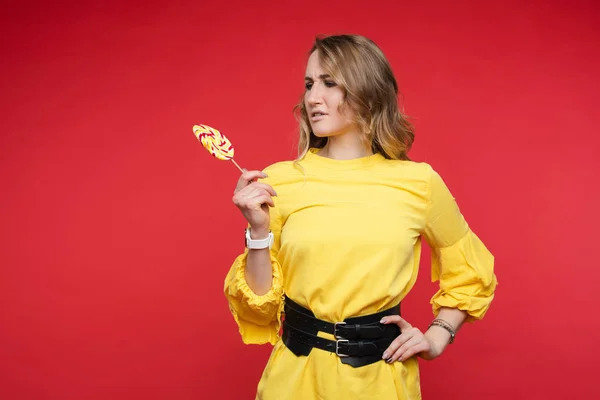 Chica confusa en vestido amarillo con cinturón mirando piruleta . —  Fotos de Stock