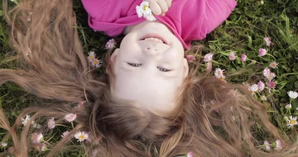 Primer plano sonriente niña linda acostada sobre hierba verde sosteniendo flores mirando a la cámara — Vídeos de Stock