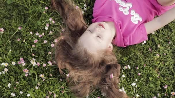 Close-up sorrindo pequena menina bonito deitado na grama verde segurando flores olhando para a câmera — Vídeo de Stock