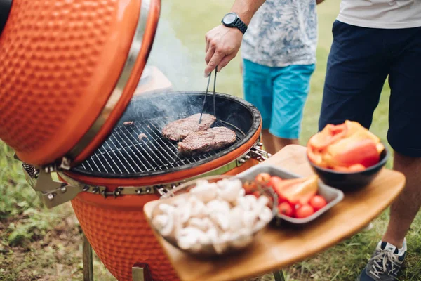 Homem irreconhecível cozinhando carne na grelha.Mesa com cogumelos fatiados e legumes. Festa de piquenique . — Fotografia de Stock