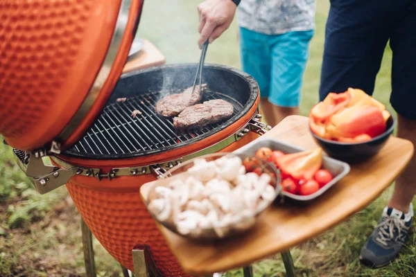 Homem irreconhecível cozinhando carne na grelha.Mesa com cogumelos fatiados e legumes. Festa de piquenique . — Fotografia de Stock