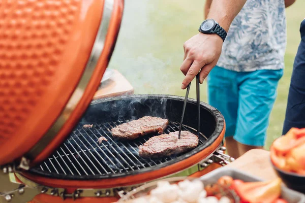 Nierozpoznawalny człowiek gotowania mięsa na grillu. Stół z plasterkami grzybów i warzyw. Letnia impreza piknikowa. — Zdjęcie stockowe