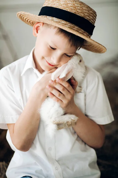 Menino de chapéu de feno mantendo bonito coelho branco e sorrindo — Fotografia de Stock