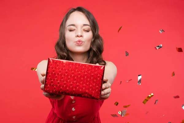 Angenehme junge Frau nimmt Geschenkschachtel küssend mit geschlossenen Augen auf rotem Studiohintergrund — Stockfoto