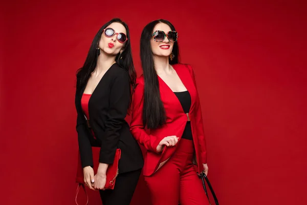 Deux jeunes mannequins féminins élégants en lunettes de soleil souriantes posant isolées au studio rouge — Photo