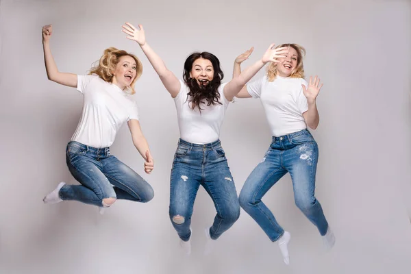 Tres amigos en ropa casual bailando y saltando. Diferentes, pero hermosas damas en jeans y camisetas blancas que se divierten y sonríen. Feliz joven rubia y morena mujeres pasar tiempo juntos . — Foto de Stock