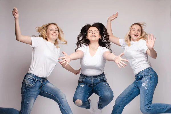 Tre amici in abiti casual che ballano e saltano. Diverse, ma belle signore in jeans e magliette bianche sempre divertenti e sorridenti. Felice giovane bionda e bruna donne trascorrere del tempo insieme . — Foto Stock