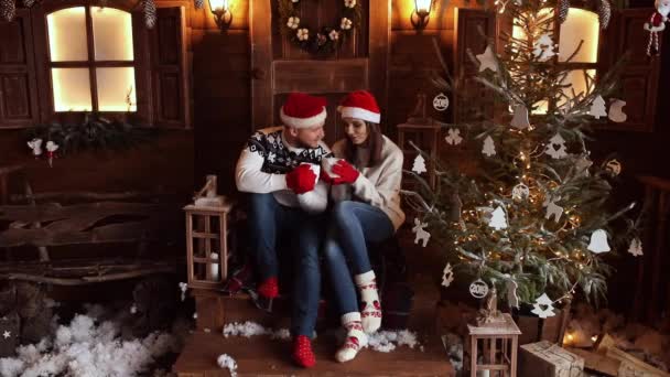 Novio y novia en sombreros de Santa sentado en el porche al lado del árbol de Navidad . — Vídeos de Stock
