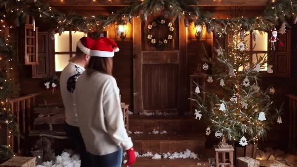 Novio y novia en sombreros de Santa sentado en el porche al lado del árbol de Navidad . — Vídeos de Stock