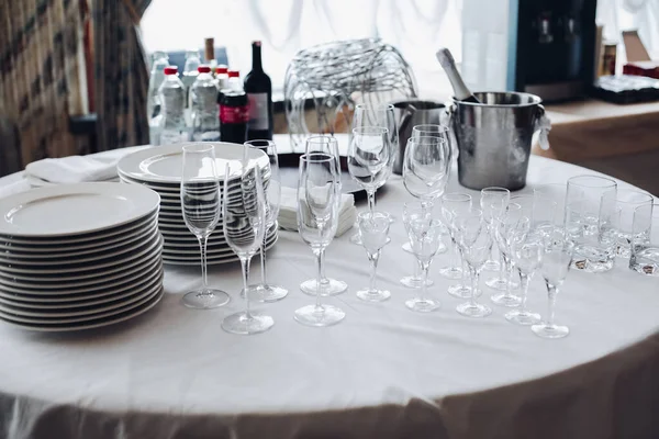 Sortiment av köksutrustning på vitt bord.Utsikt över vitt runt bord med staplar av vita tallrikar, tomma champagneflöjter och glas med flaskor med vatten och ishinkar. — Stockfoto